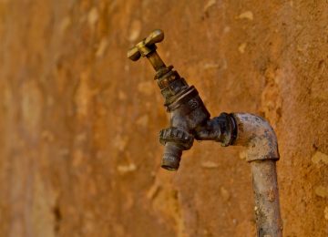 Foto de uma torneira ao lado de uma parede marrom que ilustra o post Falta de água tratada pode agravar a pandemia